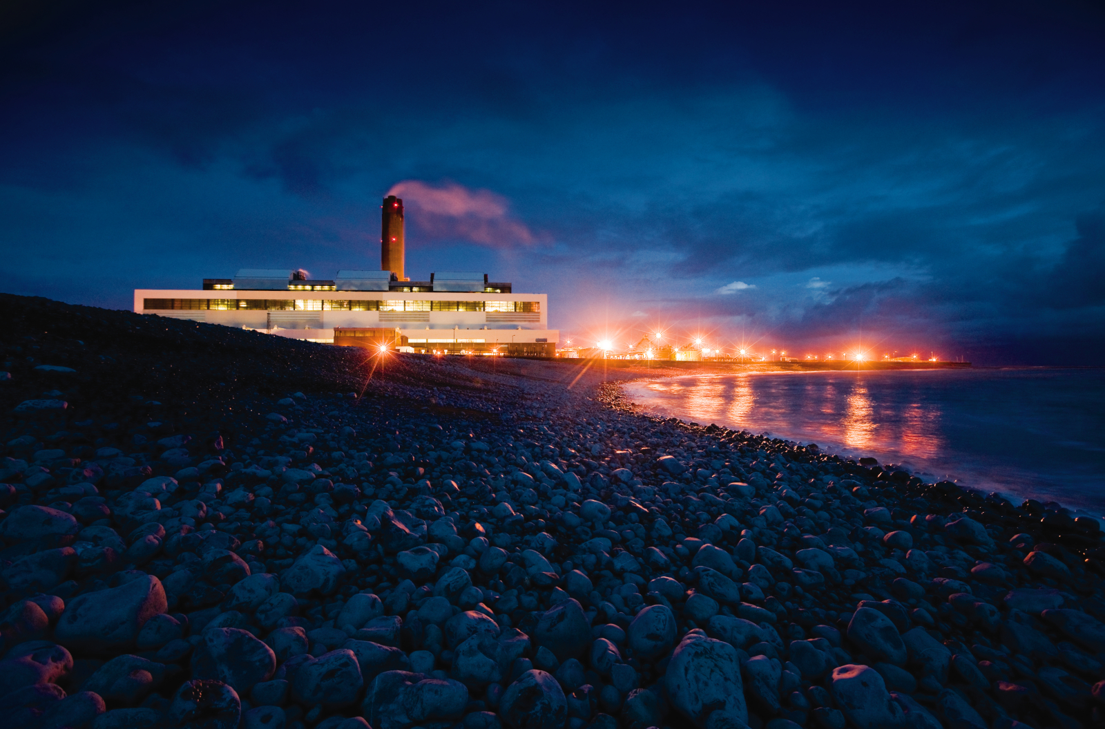 A power station by the shore at night, illuminated by lights, with rocky terrain in the foreground and a dark blue sky above.
