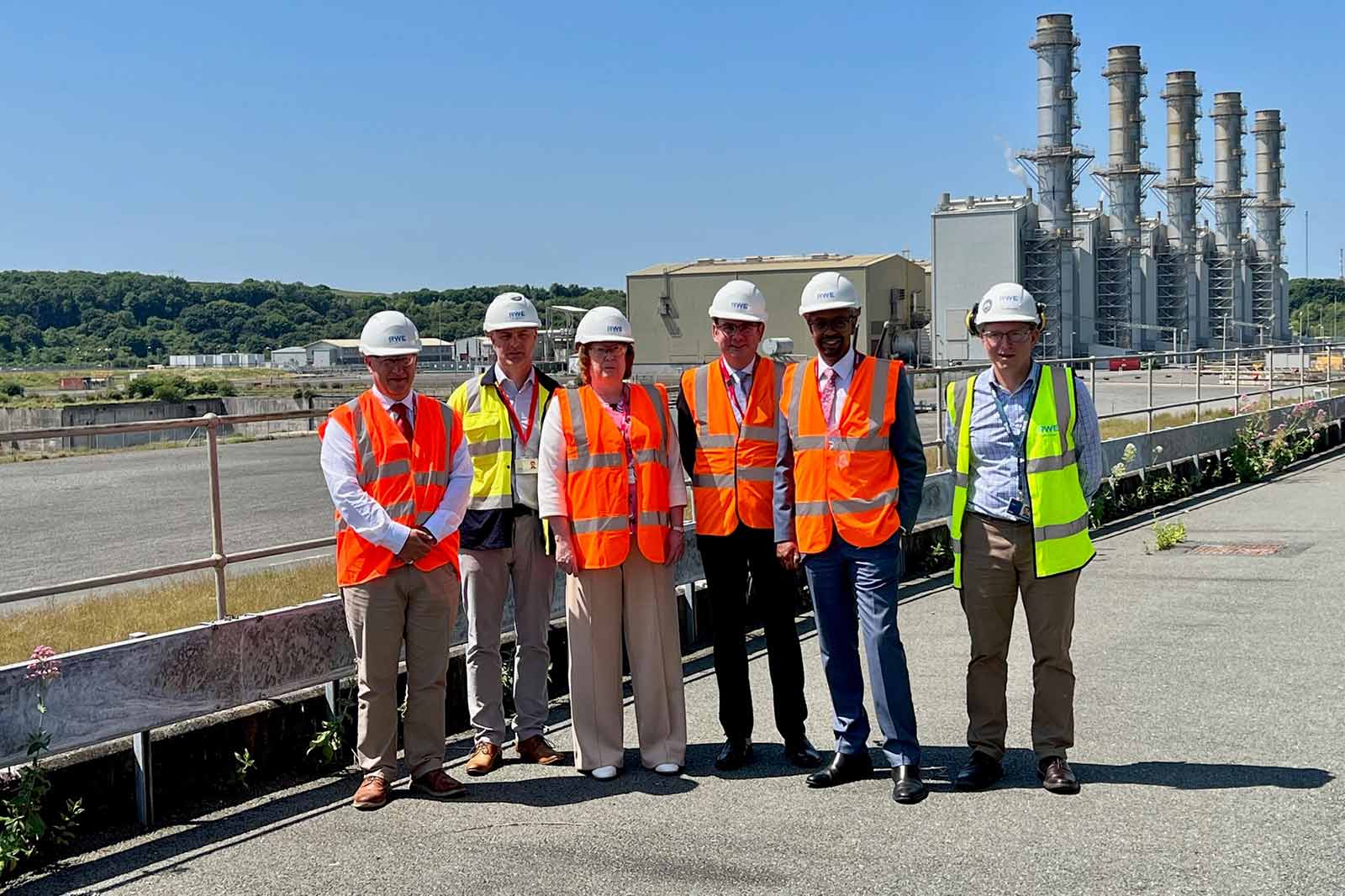 A group of six individuals in safety gear, including helmets and high-visibility vests, pose at an industrial site with a power plant backdrop.