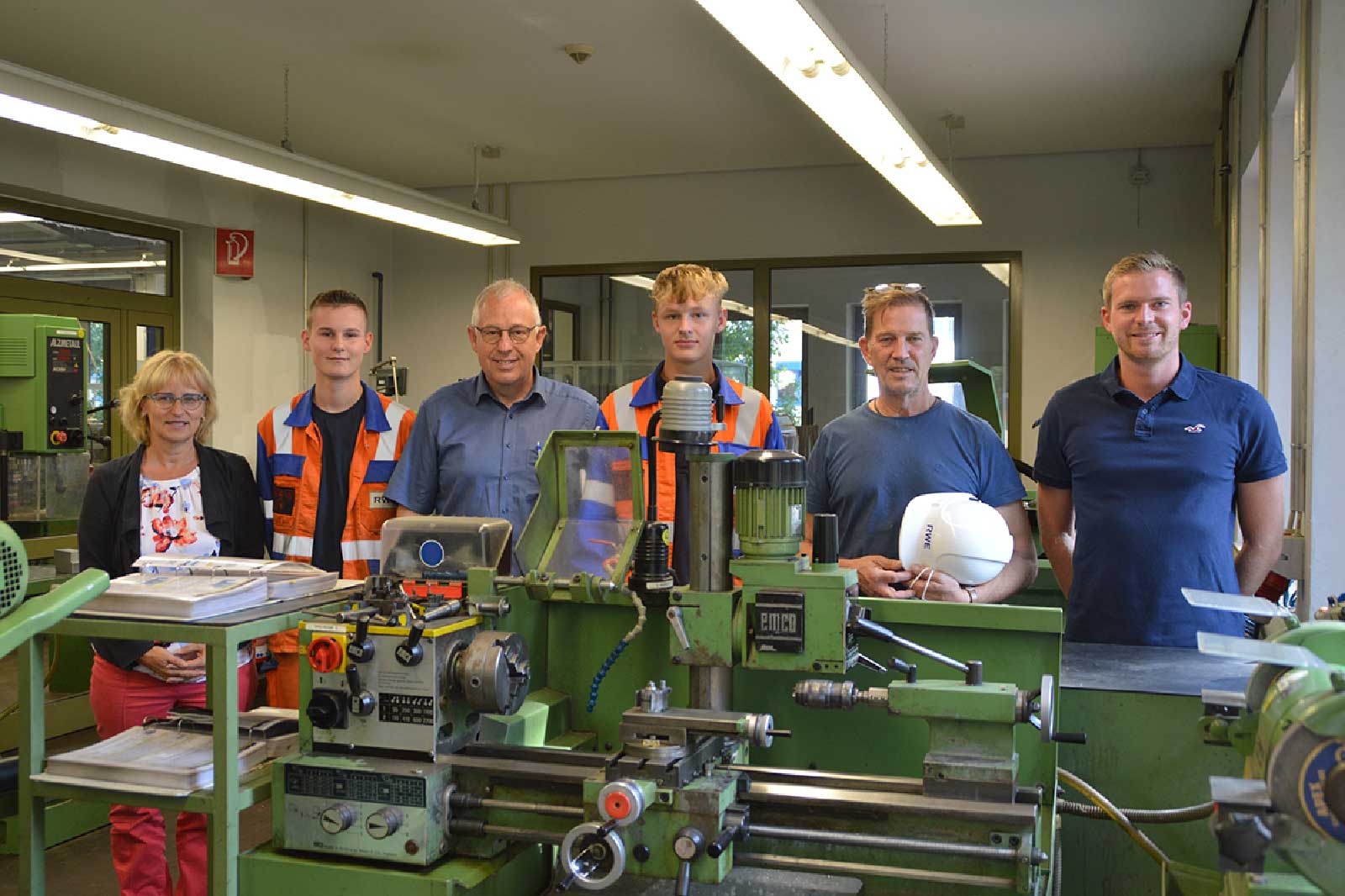 Eine Gruppe von Personen in Arbeitskleidung steht in einer Werkstatt, mit Maschinen und Werkzeugen im Vordergrund.