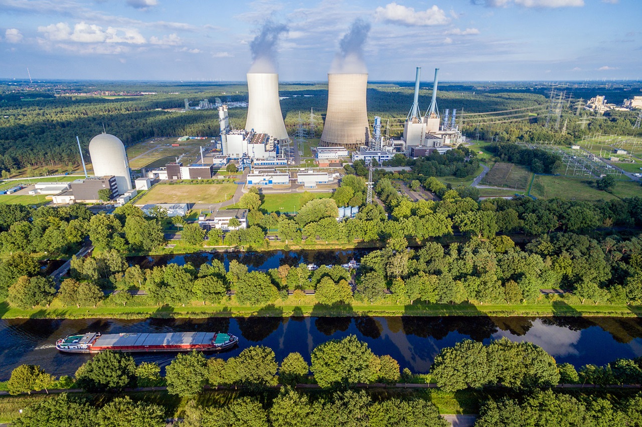 Aerial view of a power station with cooling towers and a nearby canal lined with trees.