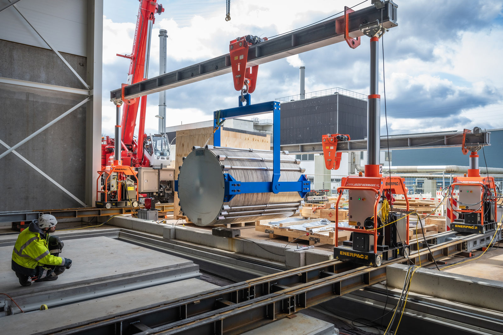 A construction site featuring a large cylindrical object being lifted by a crane, with workers and machinery visible.