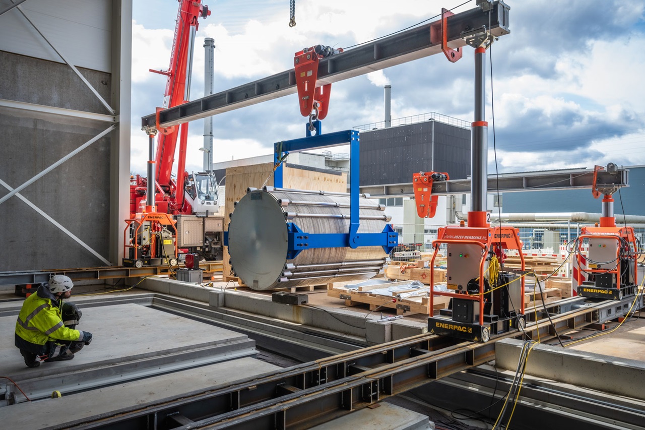 Eine Baustelle, auf der ein großes zylindrisches Objekt von einem Kran angehoben wird, mit sichtbaren Arbeitern und Maschinen.