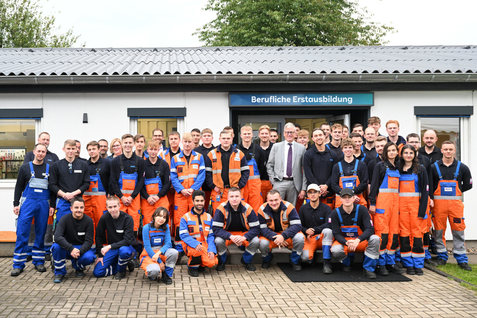 Eine Gruppe von Auszubildenden in orange-blauen Arbeitsuniformen posiert vor einem Bildungszentrum mit dem Schild 'Berufliche Erstausbildung'.
