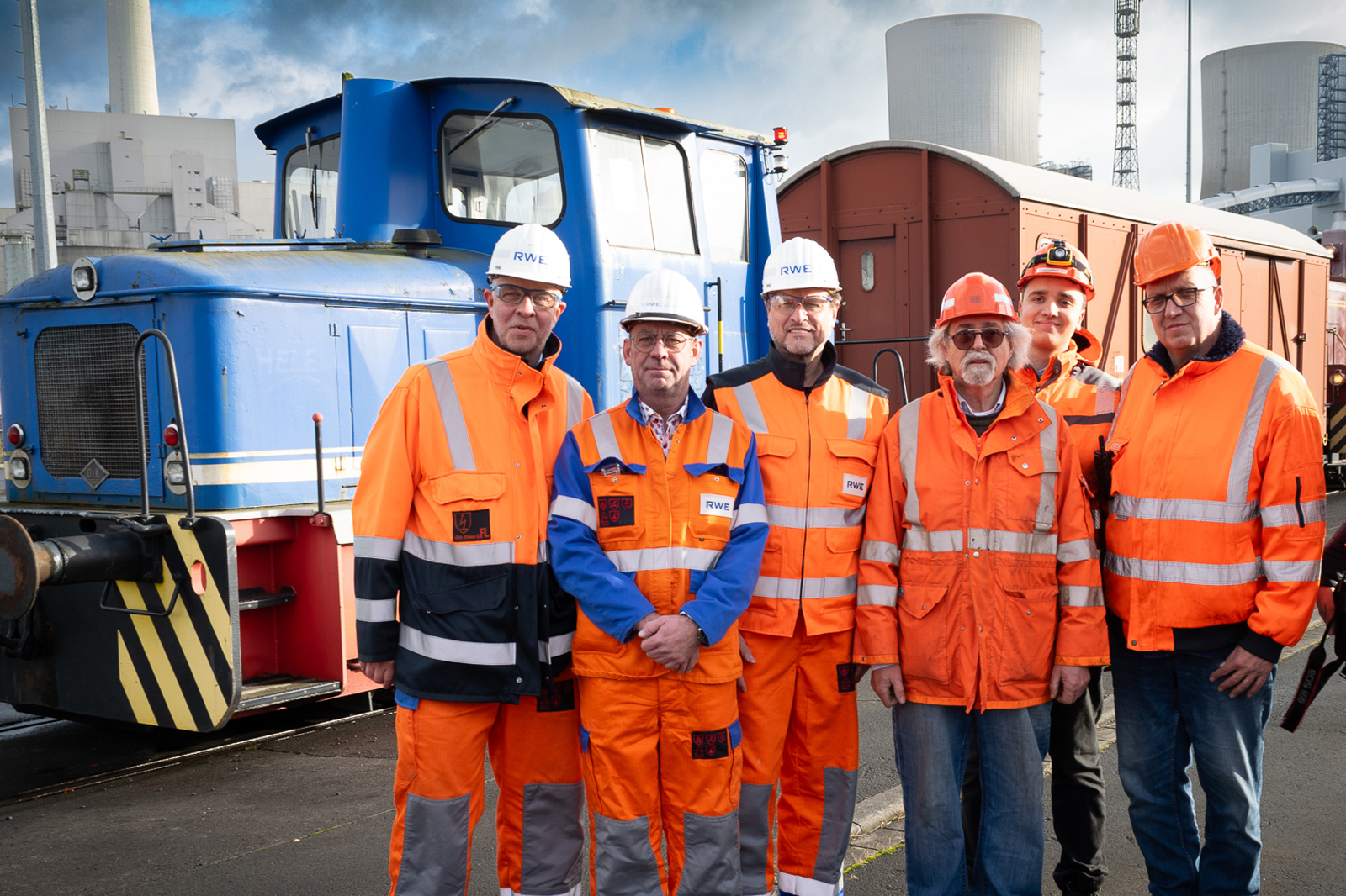 Eine Gruppe von sechs Arbeitern in hochsichtbarer orangener Kleidung steht vor einer blauen Lokomotive an einem industriellen Standort.