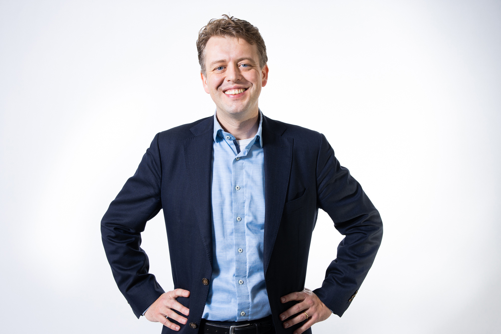 Ein Mann in einem blauen Hemd und einem dunkelblauen Blazer posiert mit Händen in den Hüften vor einem weißen Hintergrund.