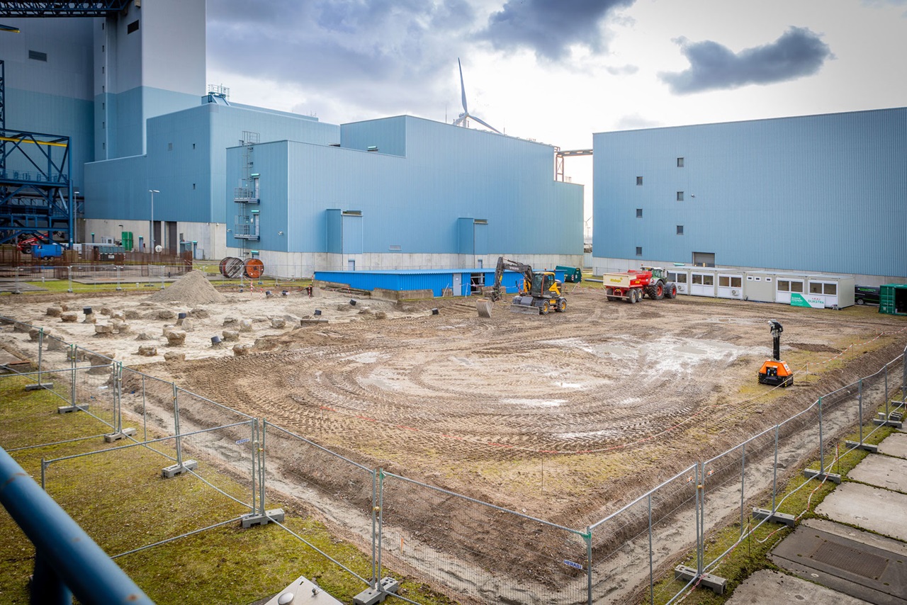 Eine industrielle Baustelle mit Maschinen und markiertem Boden, umgeben von einem Zaun und Gebäuden im Hintergrund.