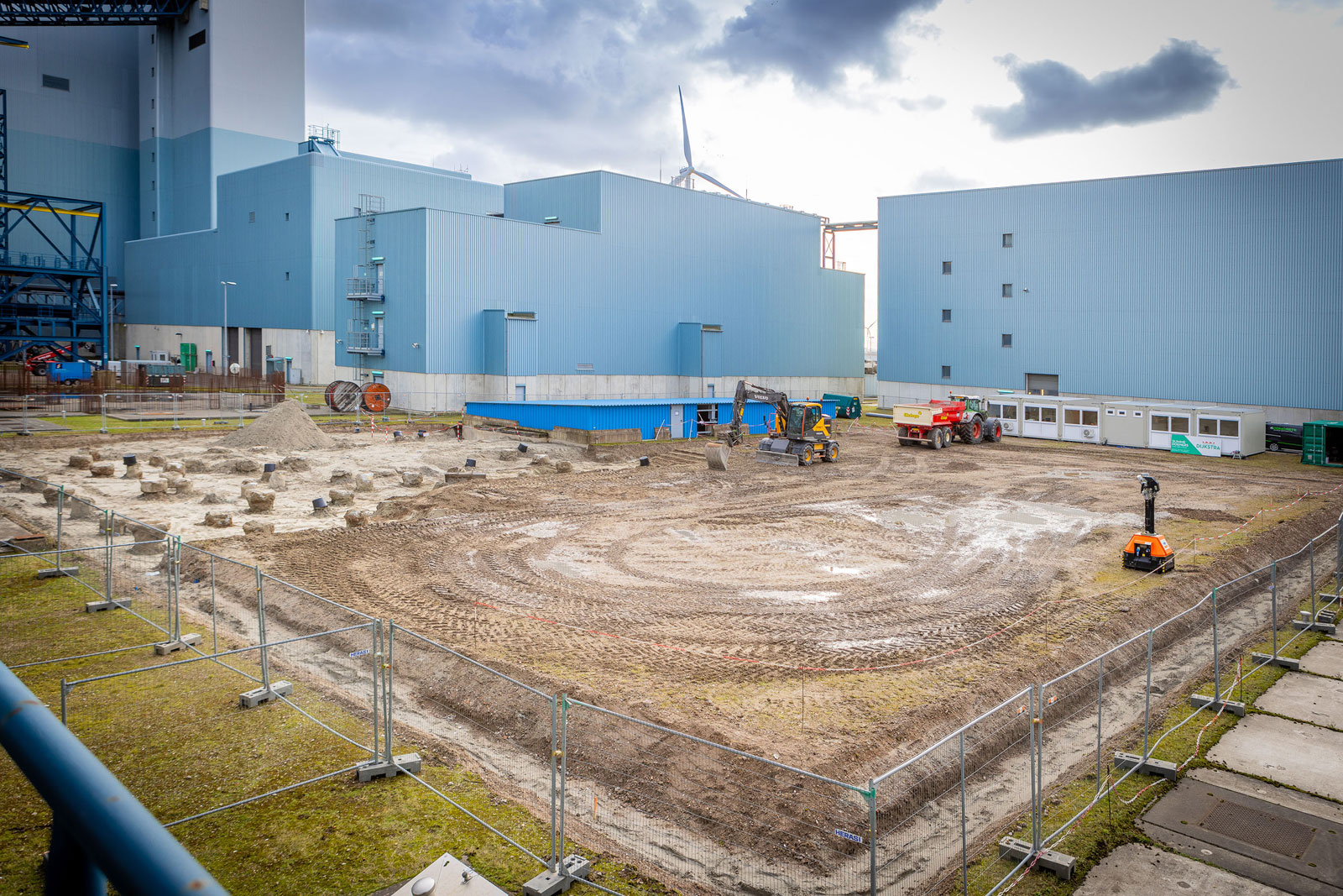 Eine industrielle Baustelle mit Maschinen und markiertem Boden, umgeben von einem Zaun und Gebäuden im Hintergrund.