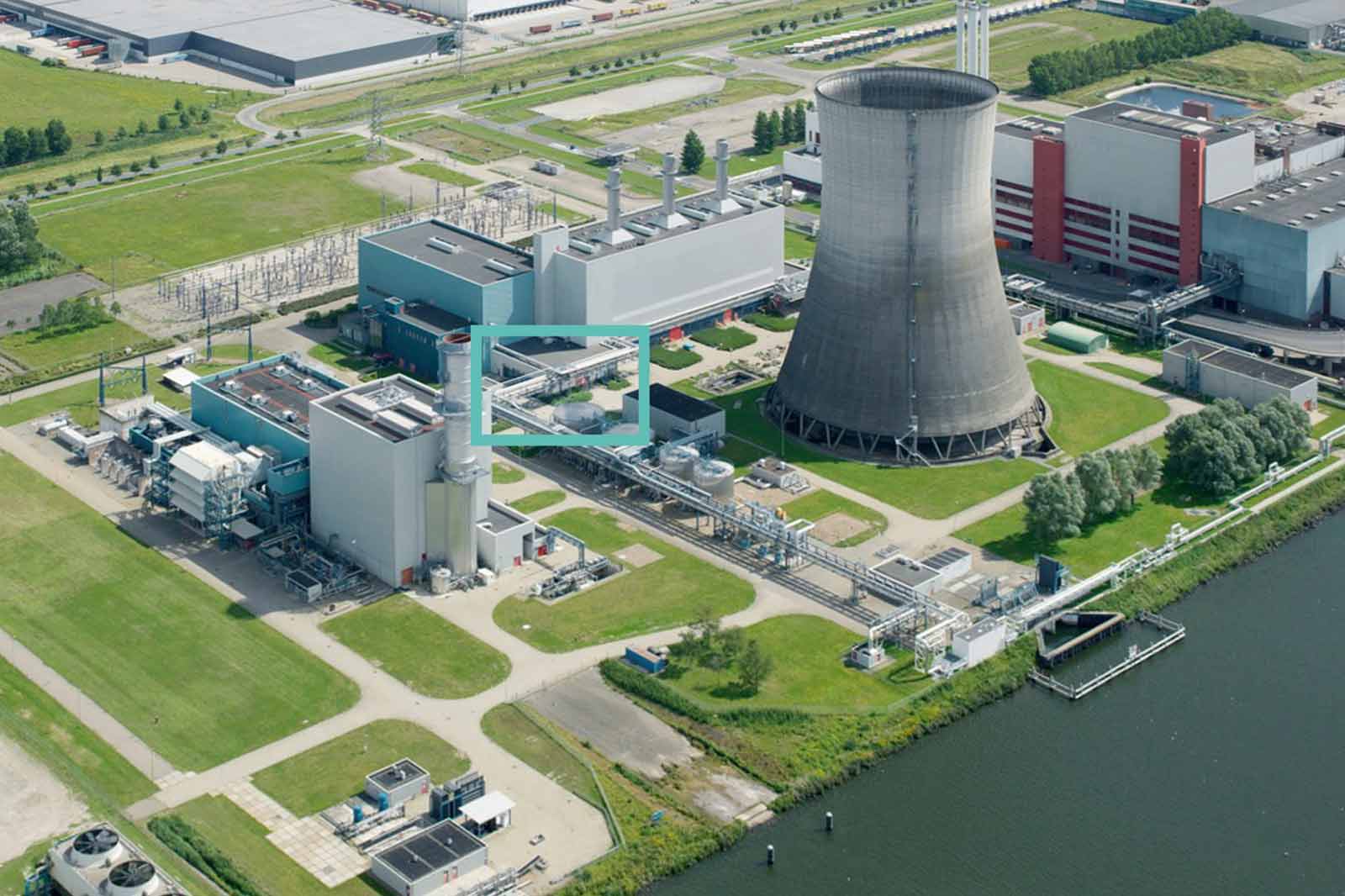 Aerial view of an industrial complex featuring a cooling tower, buildings, and power lines beside a waterway.