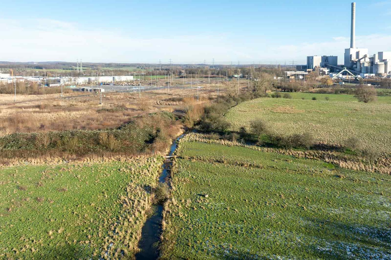 Luftaufnahme einer grünen Landschaft mit einem gewundenen Bach, im Hintergrund industrielle Gebäude unter einem klaren blauen Himmel.