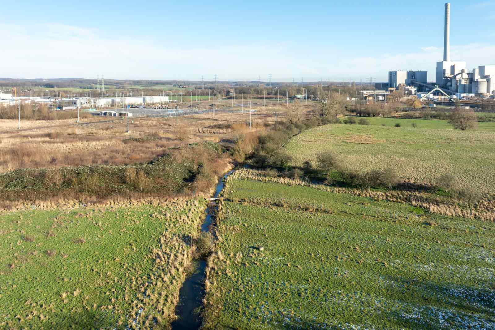 Luftaufnahme einer grünen Landschaft mit einem gewundenen Bach, im Hintergrund industrielle Gebäude unter einem klaren blauen Himmel.