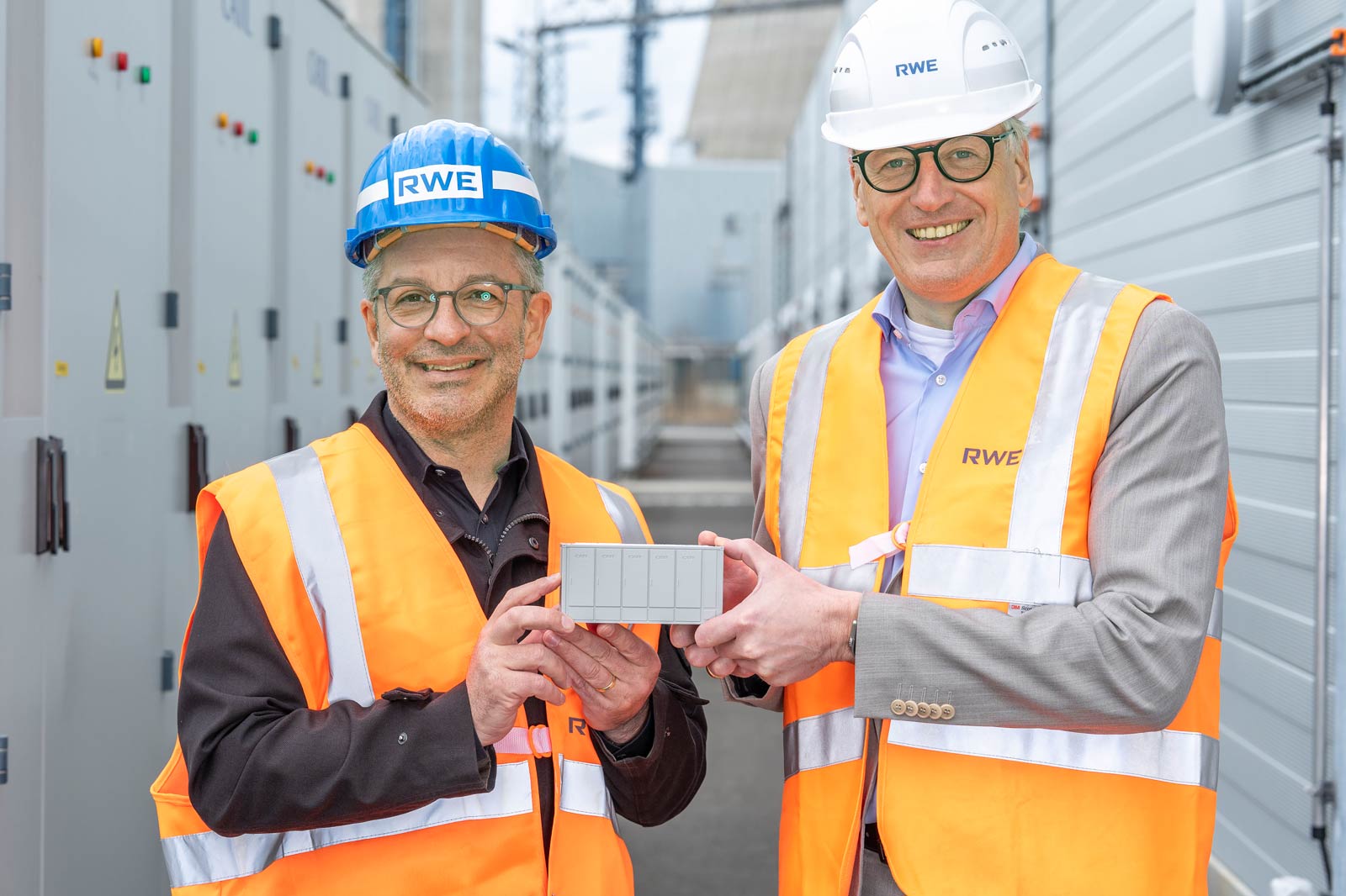Two individuals in high-visibility vests and hard hats are holding a small grey device in an industrial setting.