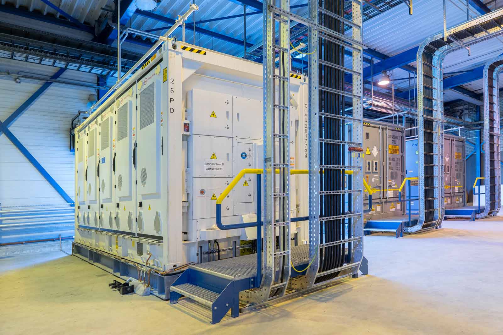 A modern battery system in an industrial setting with yellow safety steps and cable trays.