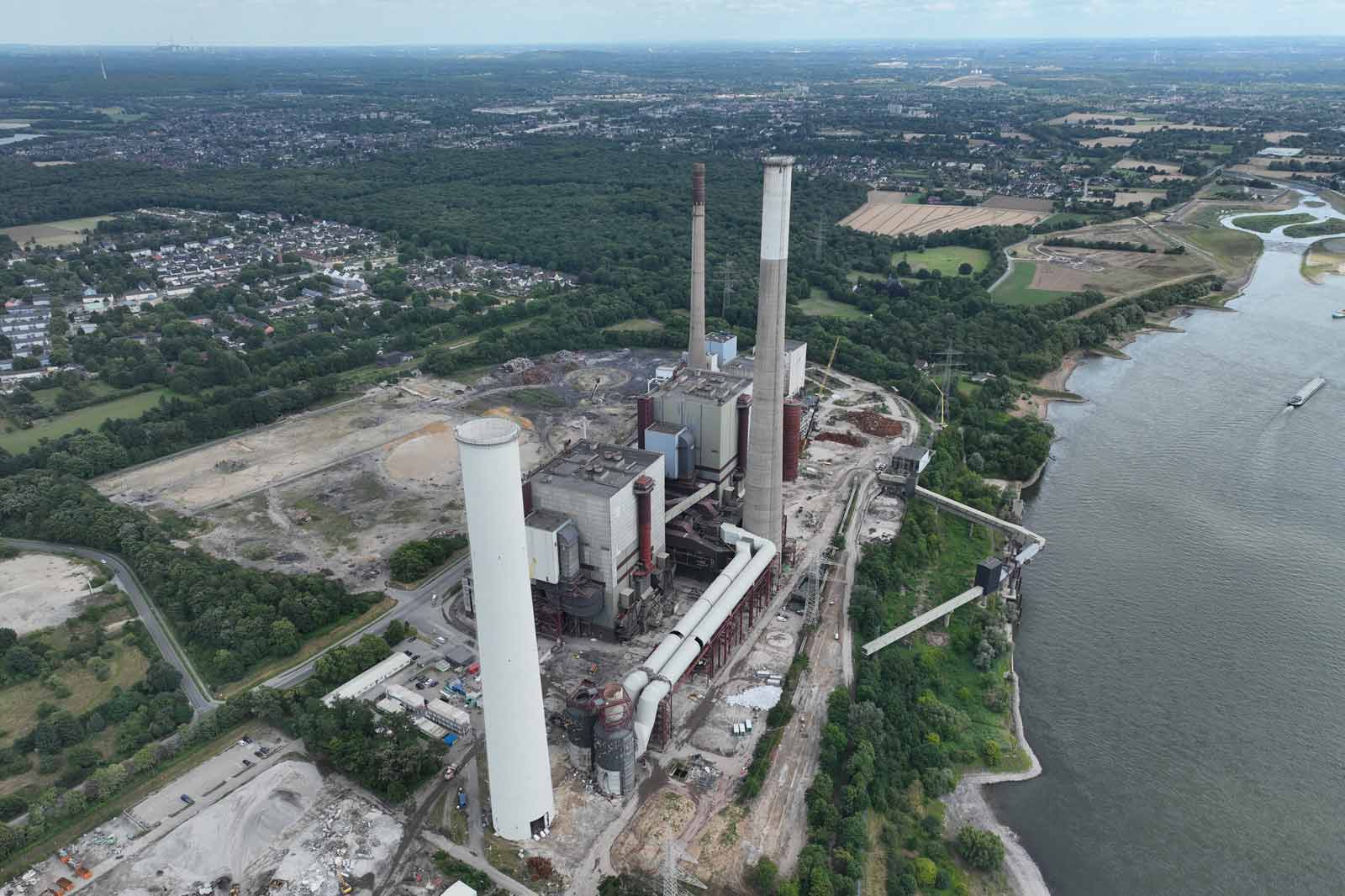Eine Luftaufnahme eines großen Kraftwerks mit Schornsteinen, in der Nähe eines Flusses und grüner Felder, umgeben von Bäumen und einem Wohngebiet.