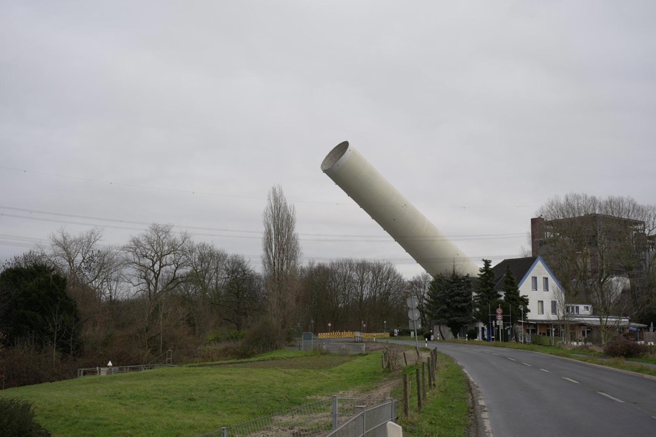 Eine geneigte, weiße zylindrische Struktur neben einer Straße, umgeben von Bäumen und Gebäuden unter einem bewölkten Himmel.