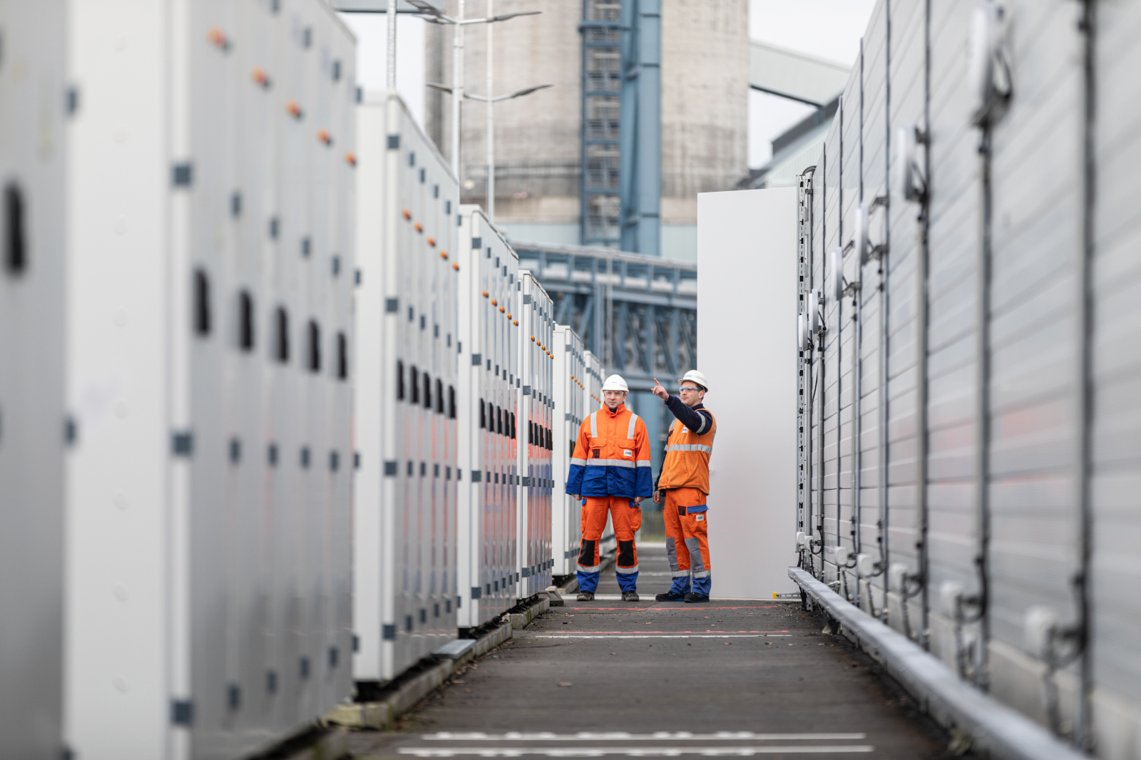 Zwei Personen in orangefarbener Arbeitskleidung stehen zwischen großen elektrischen Schränken auf einem Industriegelände.