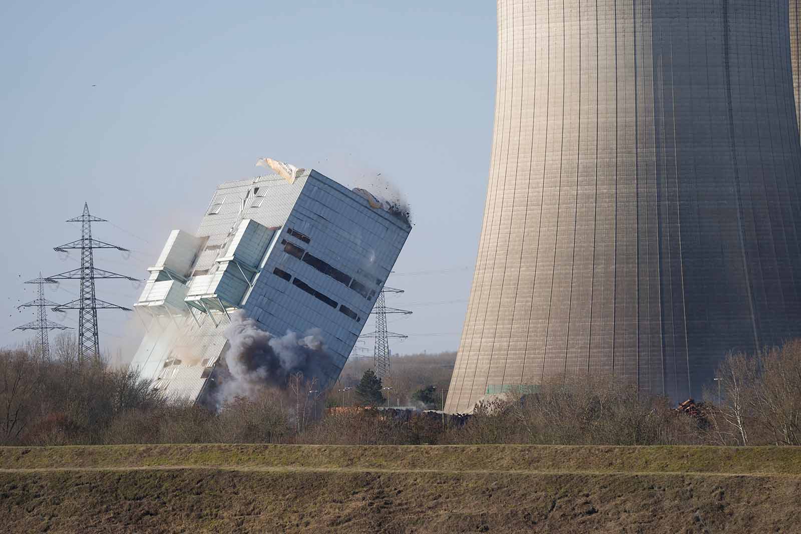 Ein großes Industriegebäude stürzt ein, es entstehen Staub- und Rauchwolken; ein hoher Kühlturm, Strommasten und kahle Bäume sind sichtbar.