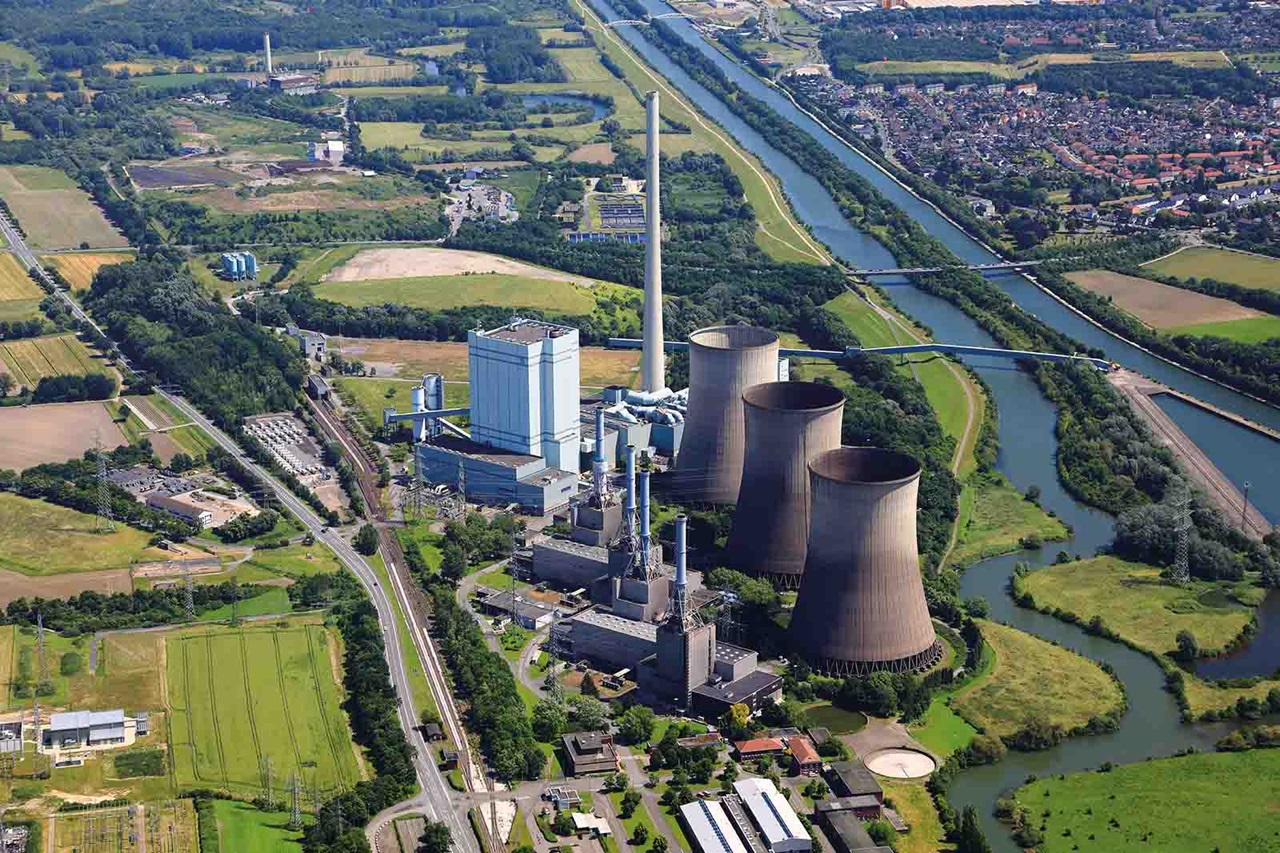 Aerial view of an industrial complex dominated by four large concrete cooling towers and a tall chimney beside a winding blue river and green fields.