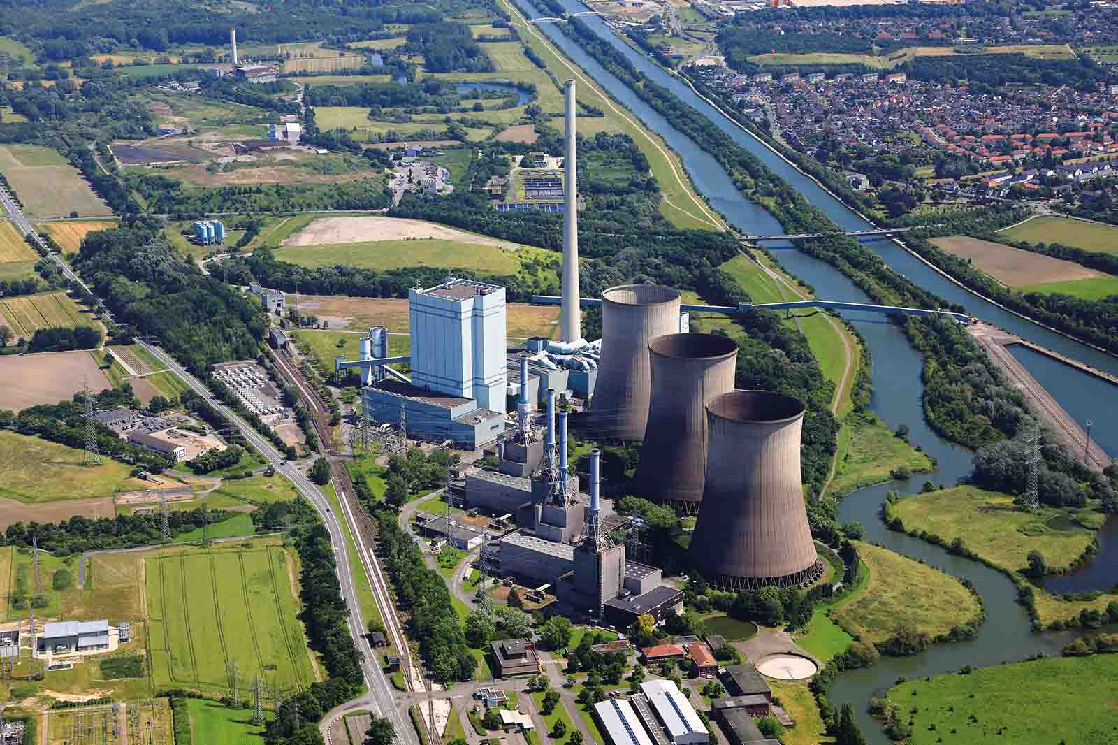 Luftaufnahme eines Industriekomplexes mit vier großen Betonkühltürmen und einem hohen Schornstein neben einem blauen Fluss, grünen Feldern und einer Kleinstadt.