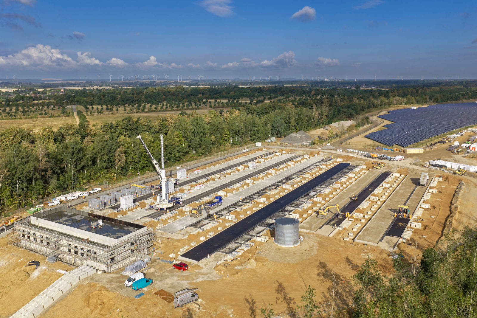 Luftaufnahme einer weitläufigen Baustelle für eine Solaranlage mit Reihen dunkler Solarmodule, einem Turmdrehkran, einem runden Tank und umgebenem Wald.