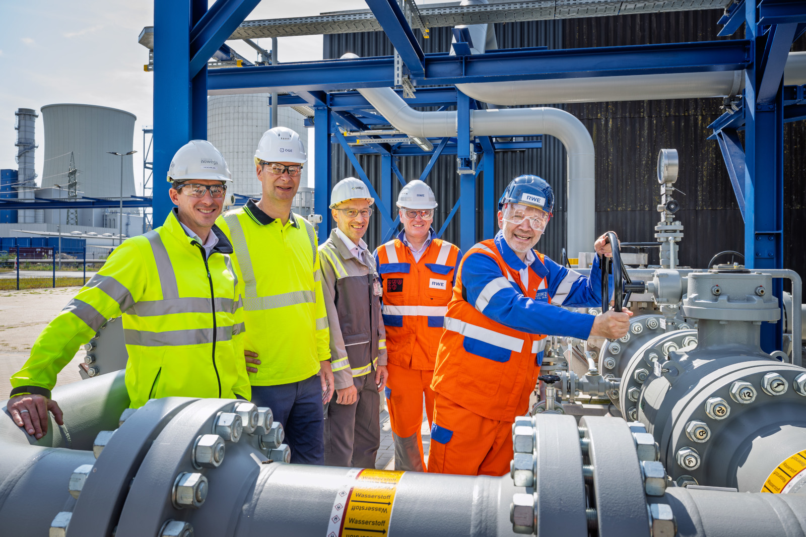 Five workers in helmets and high-visibility clothing stand by large metal pipes while one in orange coveralls turns a grey valve, smiling.