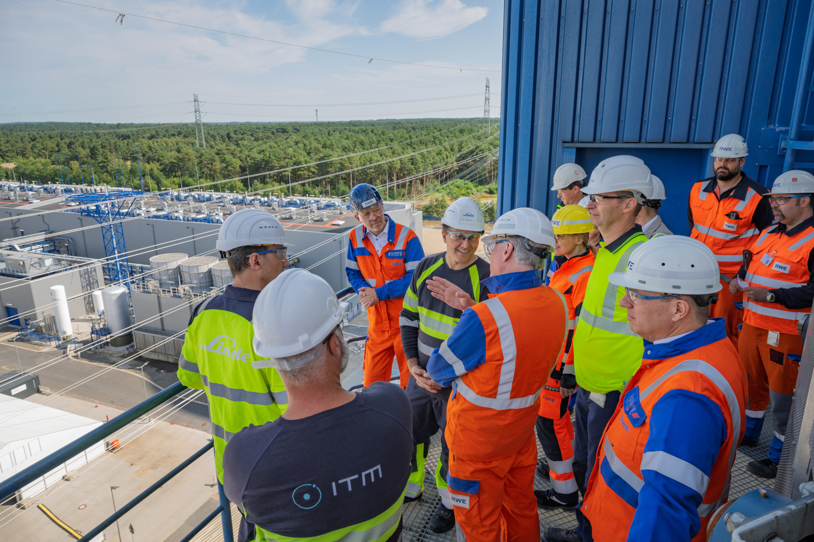 Workers in white and yellow hard hats and orange and blue high-visibility coveralls stand on a metal platform at an industrial plant, one shirt reads "ITM".