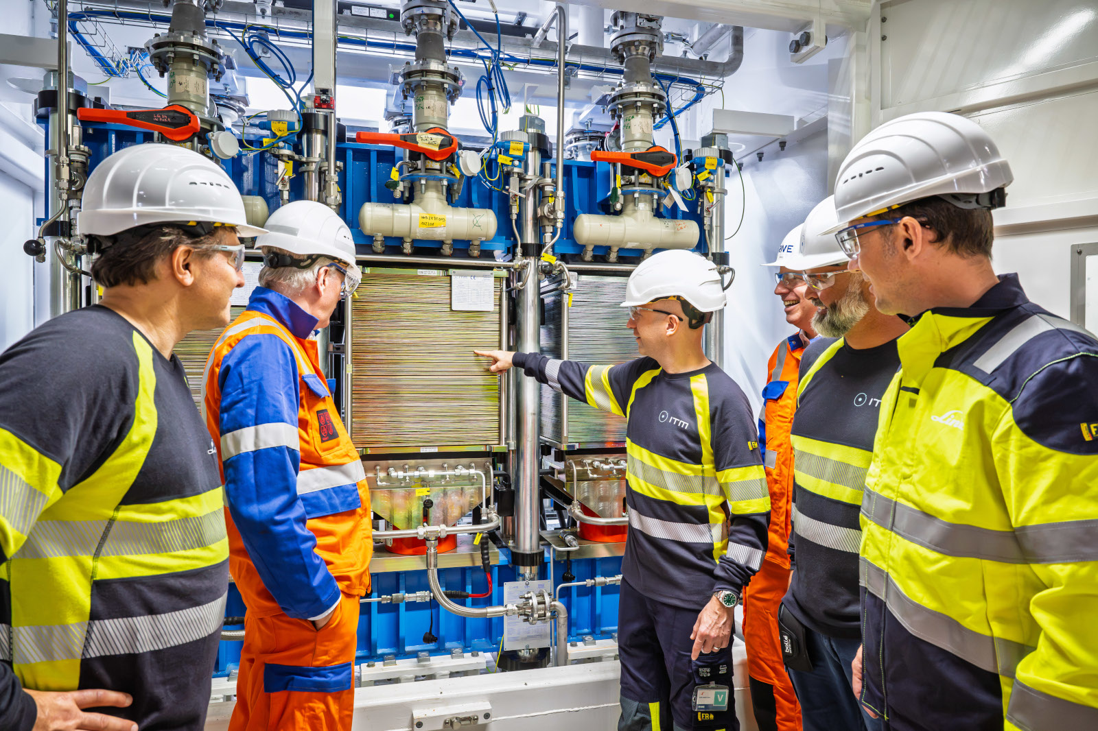 Six workers in white helmets and yellow and orange high-visibility clothing stand around stainless-steel industrial machinery while one person points at a control panel.