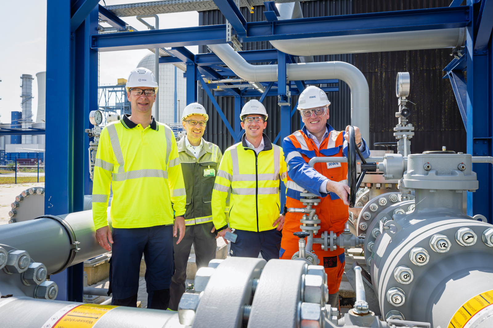 Four smiling workers in white helmets and high-visibility jackets, one in orange coveralls, stand amid blue steel framing and large gray pipes.