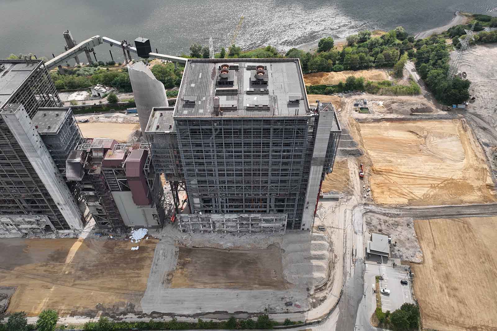 Ein großer, mehrstöckiger Industriekomplex mit sichtbarer Stahlrahmenfassade und Maschinen auf dem Dach steht neben kahlen, sandfarbenen Bauflächen am Ufer eines Flusses.