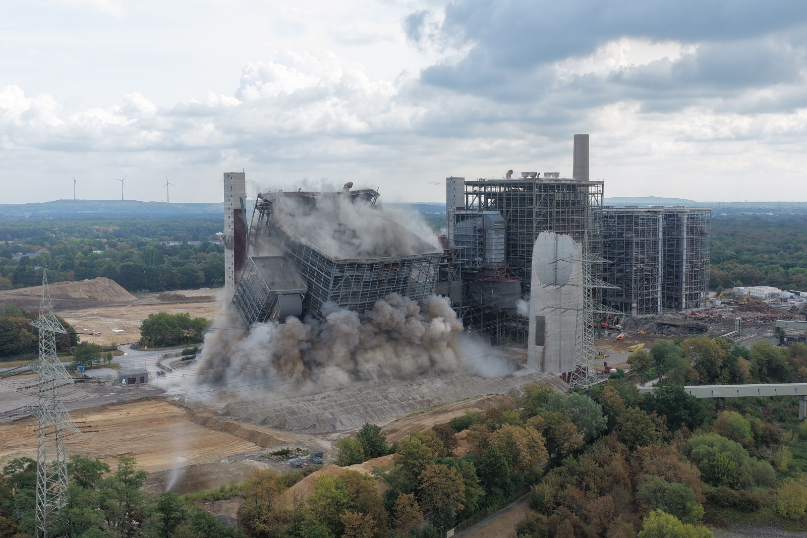 Ein großes graues Industriegebäude stürzt in sich zusammen, dichte Staub- und Rauchwolken steigen auf und umgeben freiliegende Stahl- und Betonstrukturen unter bewölktem Himmel.