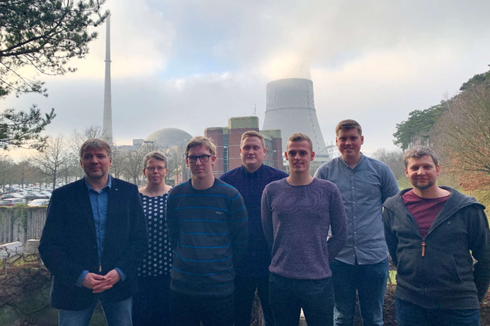 Eine Gruppe von sieben Personen steht im Freien in der Nähe eines Kraftwerks mit Kühltürmen und einem Schornstein im Hintergrund.