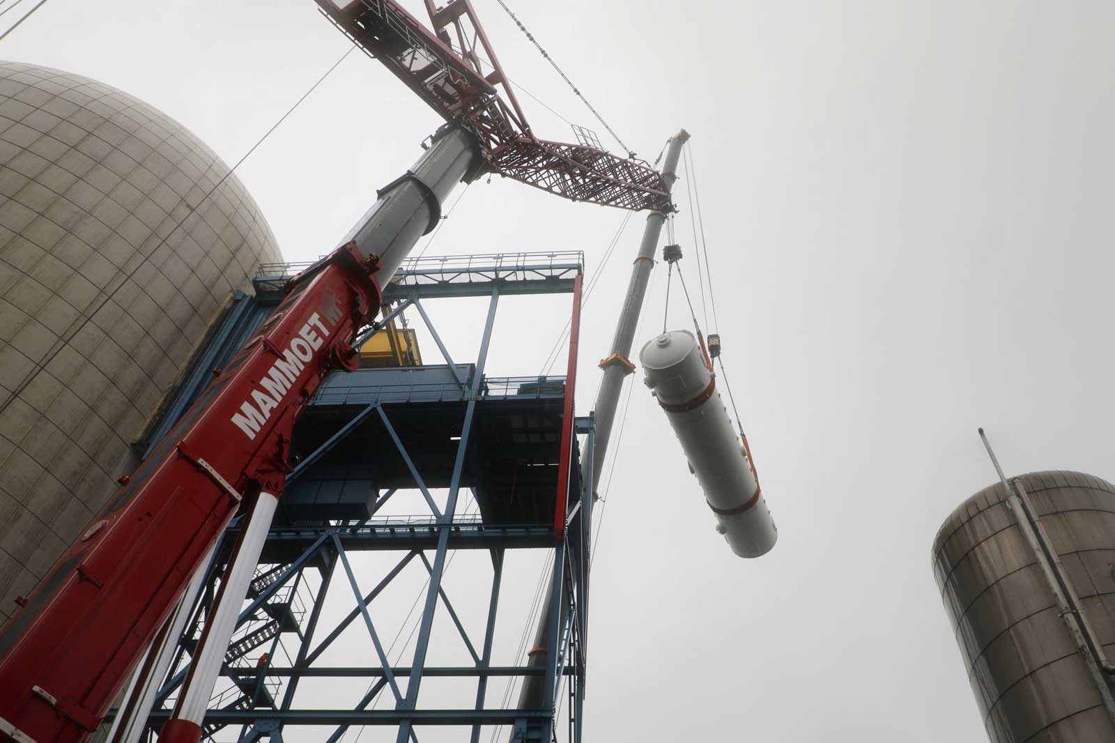 Ein Kran hebt ein zylindrisches Objekt an einem Industriegelände unter bewölkten Himmel.