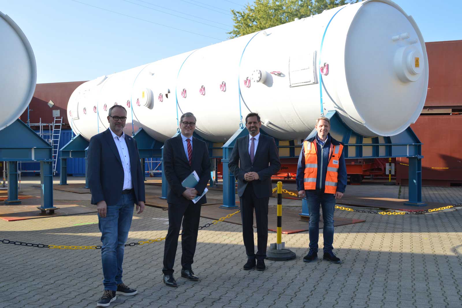 Vier Personen in formeller Kleidung stehen in der Nähe großer zylindrischer Industrieanlagen in einem Betrieb.