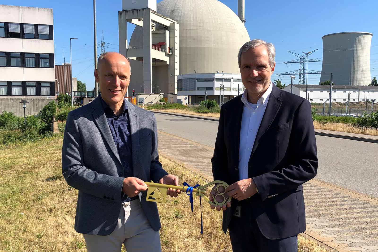 Zwei Männer stehen vor einem Kernkraftwerk und halten einen symbolischen Schlüssel, mit einem Reaktordom und Kühltürmen im Hintergrund.
