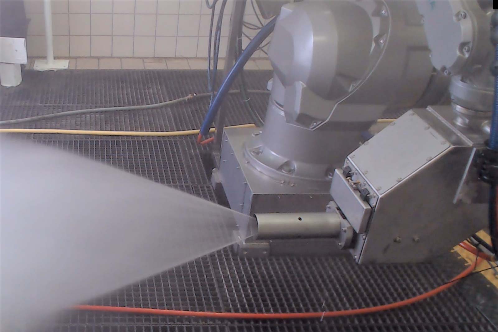 Ein Roboterarm sprüht Wasser in einer kontrollierten Umgebung, mit sichtbaren Schläuchen und einem Gitterboden.