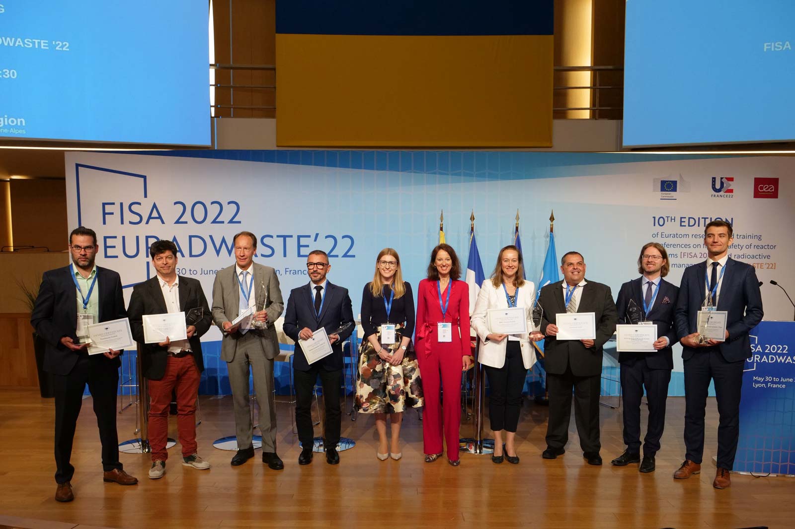 Teilnehmer der FISA 2022-Konferenz, die auf der Bühne Zertifikate halten, mit Fahnen im Hintergrund.