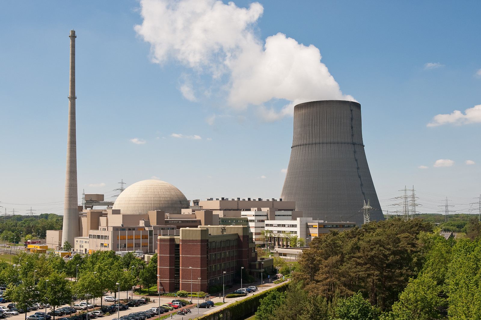 Ein Kernkraftwerk mit einem Kühlturm, einer gewölbten Struktur und einem hohen Schornstein vor blauem Himmel und Grün.
