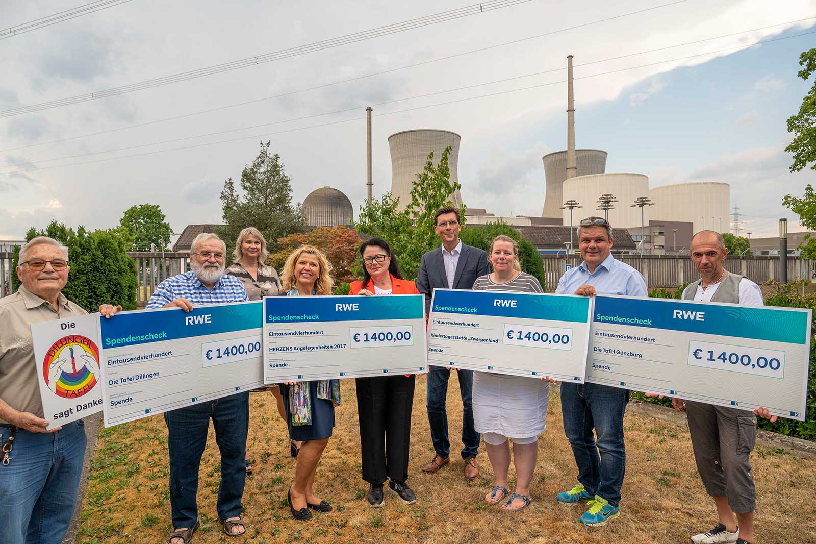 Eine Gruppe hält übergroße Spendenchecks für verschiedene Wohltätigkeitsorganisationen im Freien, mit Industriegebäuden im Hintergrund.