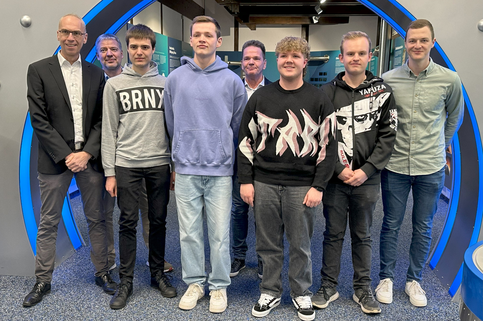 Gruppe von acht Personen, die in einem modernen Ausstellungsraum mit blauen Akzenten zusammen posieren.