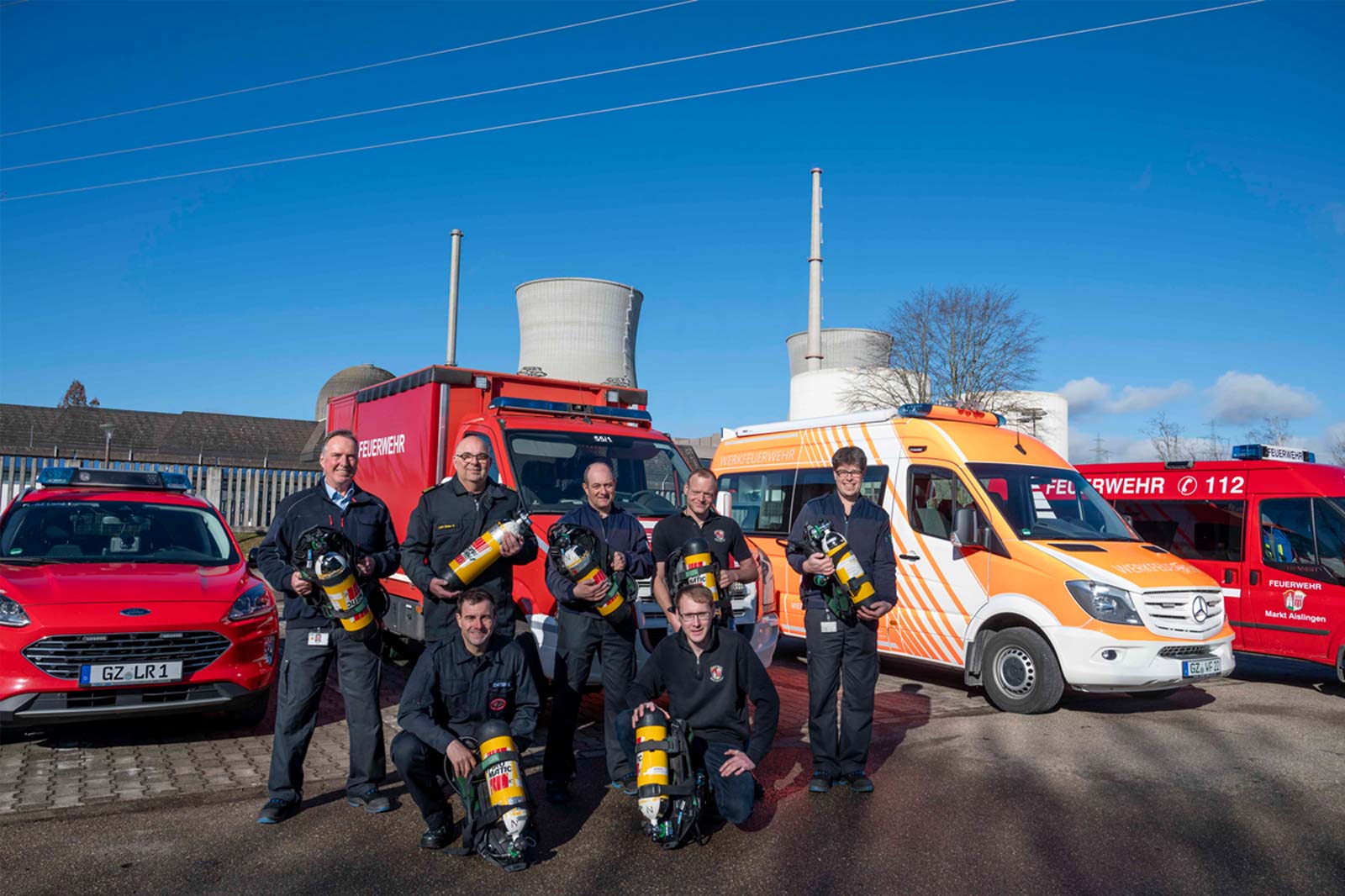 Feuerwehrleute posieren mit Ausrüstung vor Einsatzfahrzeugen und Kühltürmen an einem klaren Tag.