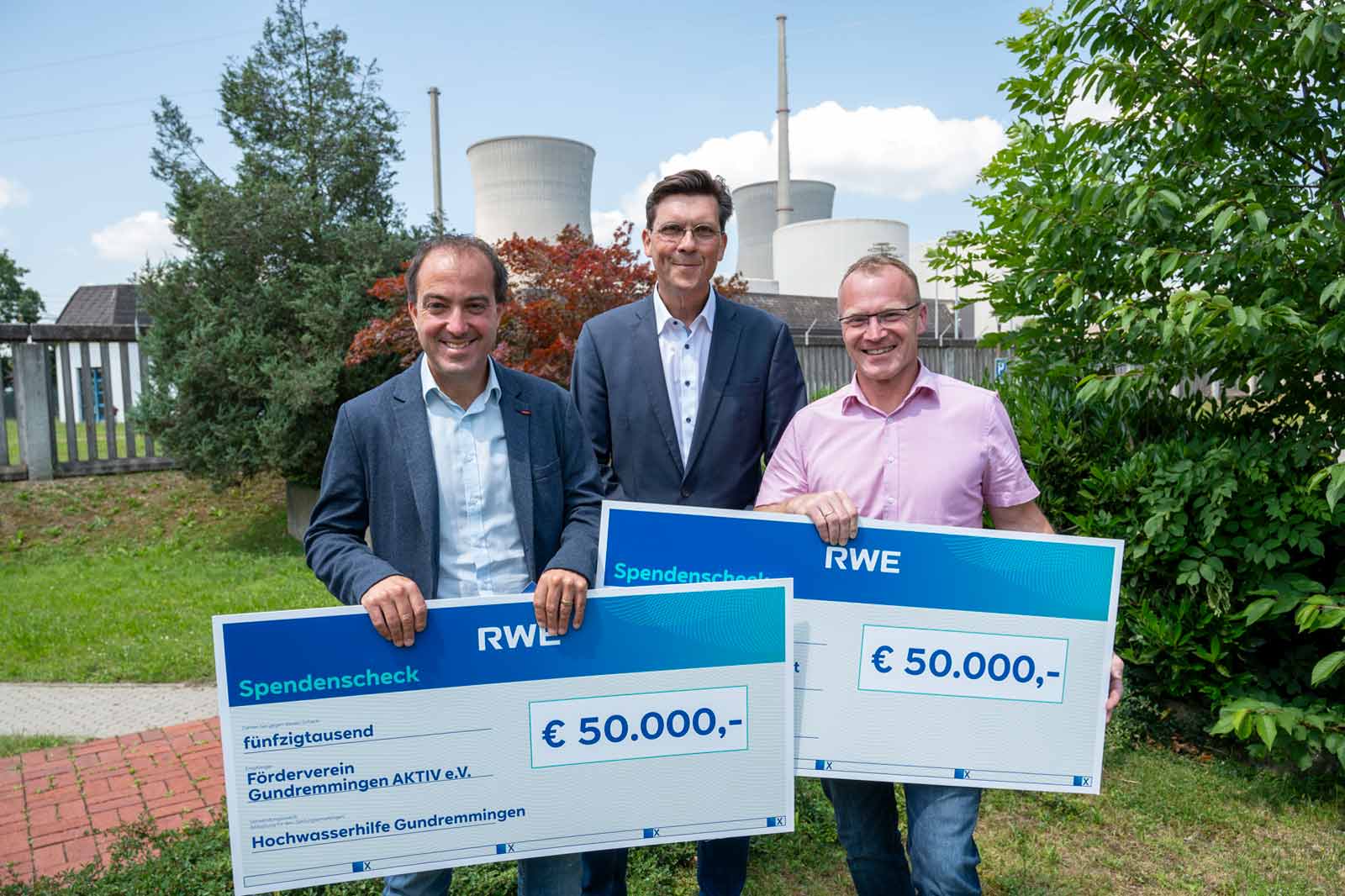 Drei Personen halten große Spendenchecks vor einem Kraftwerk, die 50.000 € für die Gemeinschaftsunterstützung zeigen.