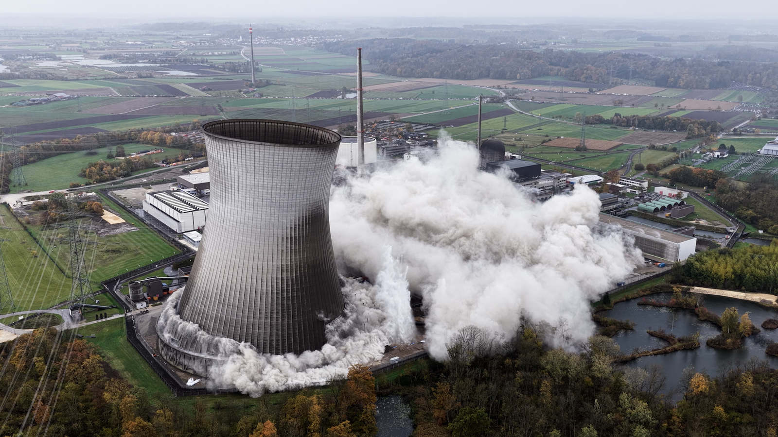 Ein Kühlturm des Kraftwerks Gundremmingen bricht zusammen und setzt eine große Wolke aus Staub und Rauch frei, umgeben von grünen Feldern und Stromleitungen.