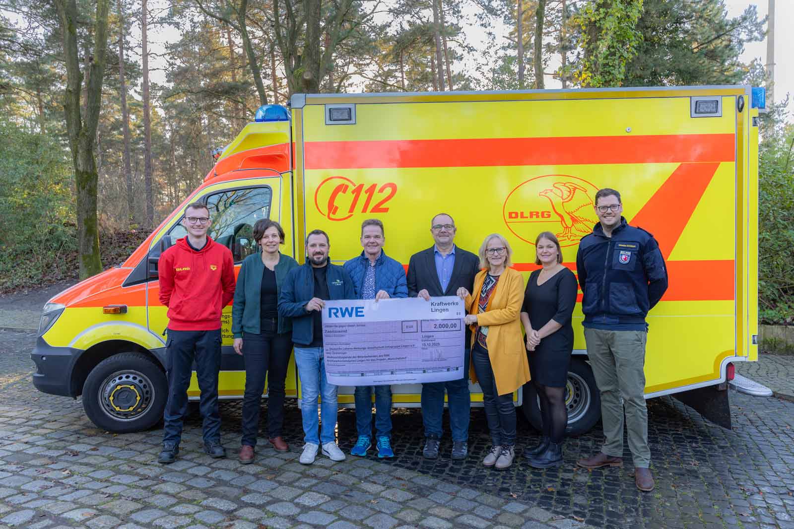 Eine Gruppe posiert vor einem hellen Einsatzfahrzeug und hält einen großen Scheck für die Wohltätigkeit.