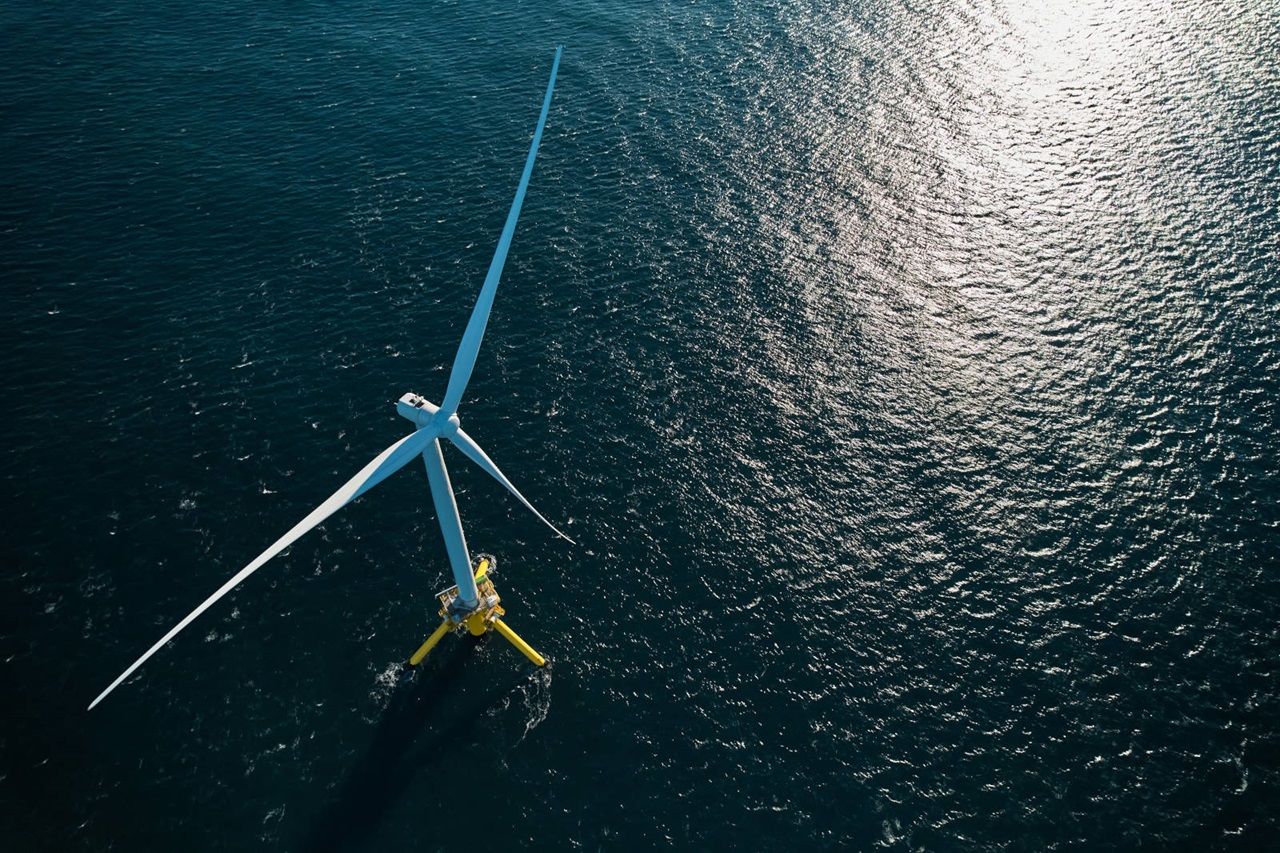 An aerial view of a large wind turbine on a blue sea, with shimmering waves reflecting sunlight.