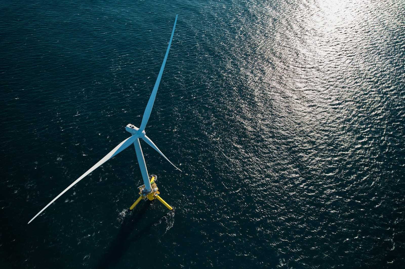 An aerial view of a large wind turbine on a blue sea, with shimmering waves reflecting sunlight.