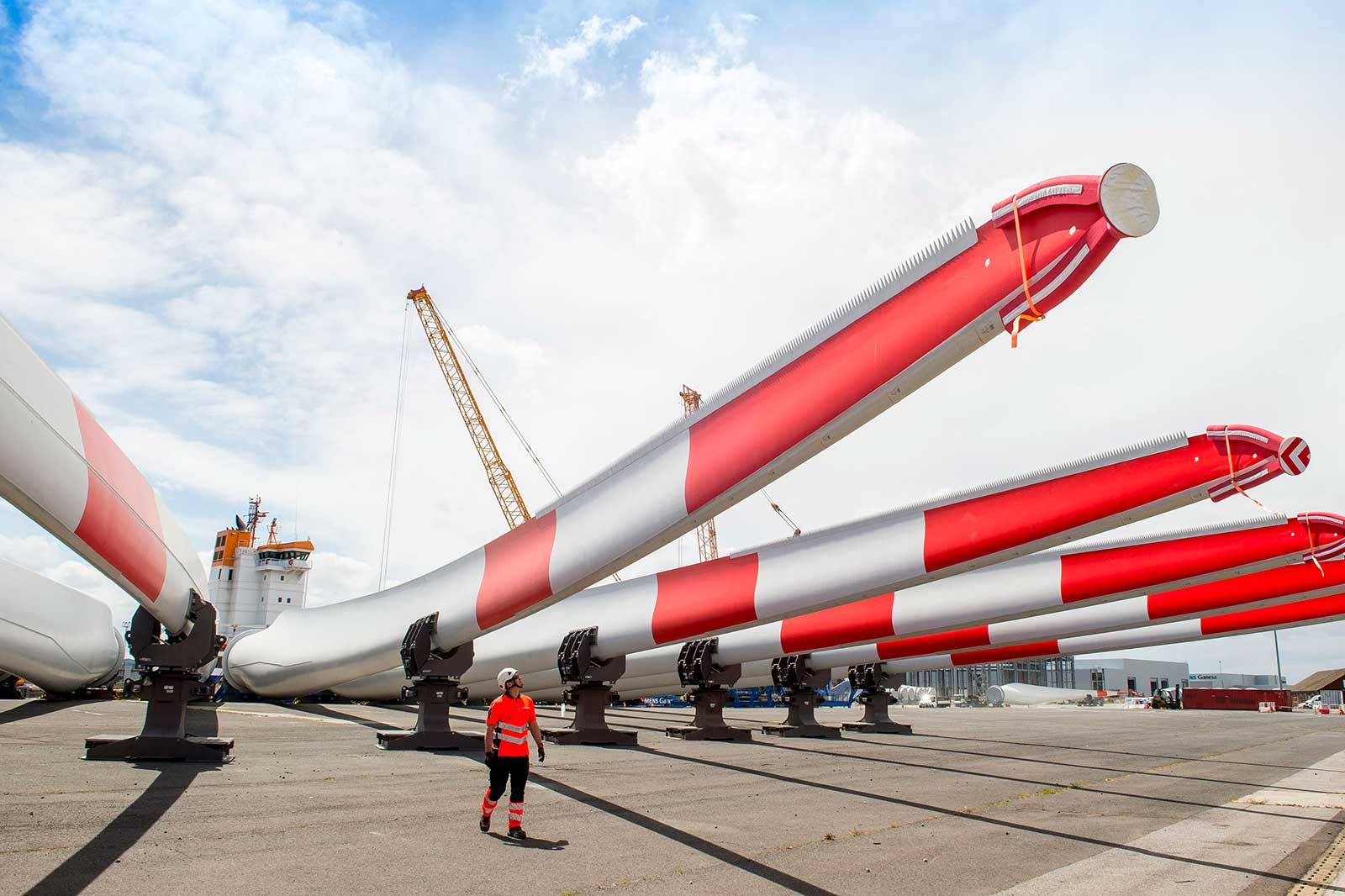 Ein Arbeiter mit Helm und Warnkleidung geht zwischen großen Windkraftanlagenblättern auf einem Dock.