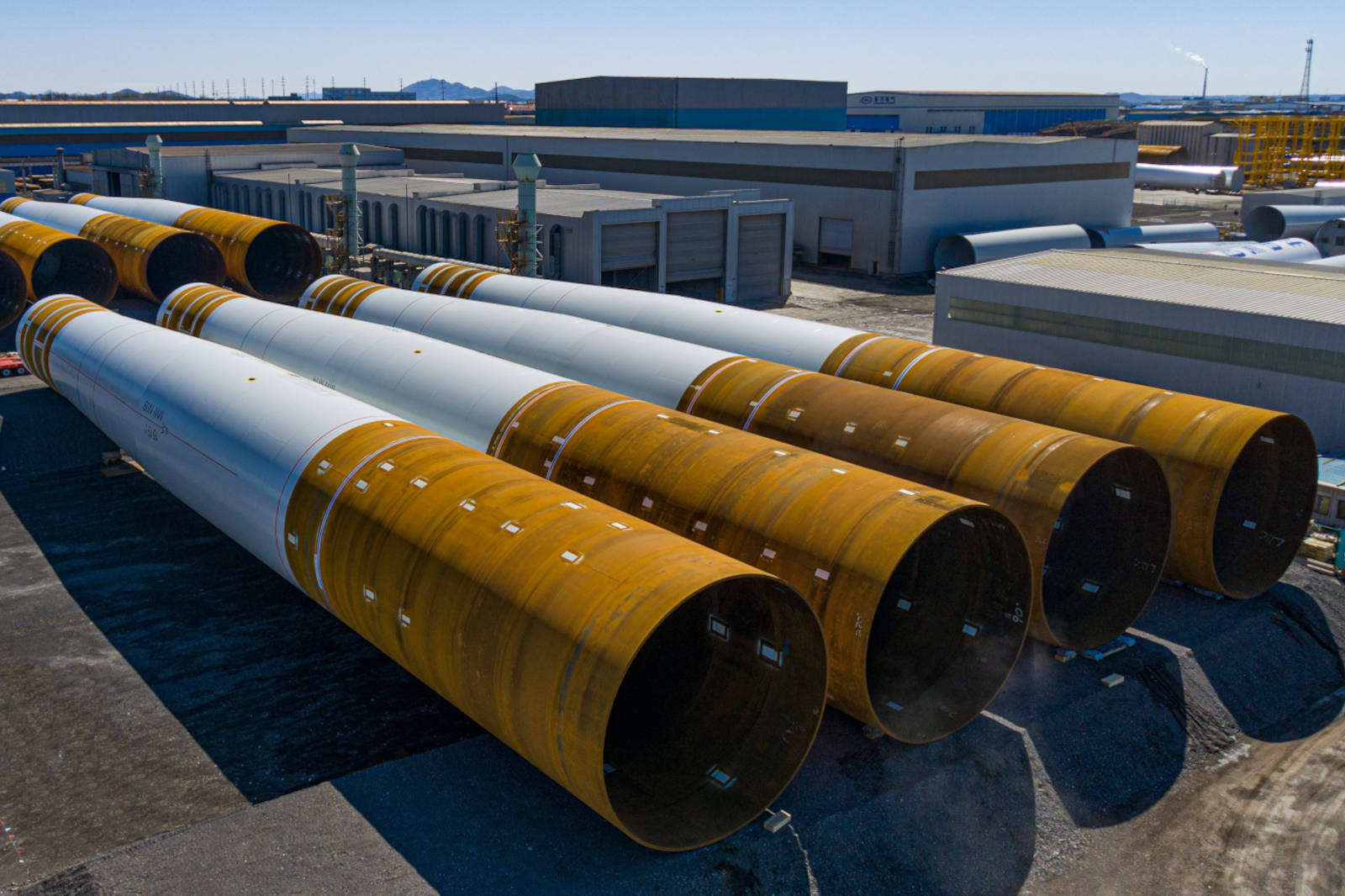Several large cylindrical structures with rusted ends are arranged on a construction site, surrounded by industrial buildings.