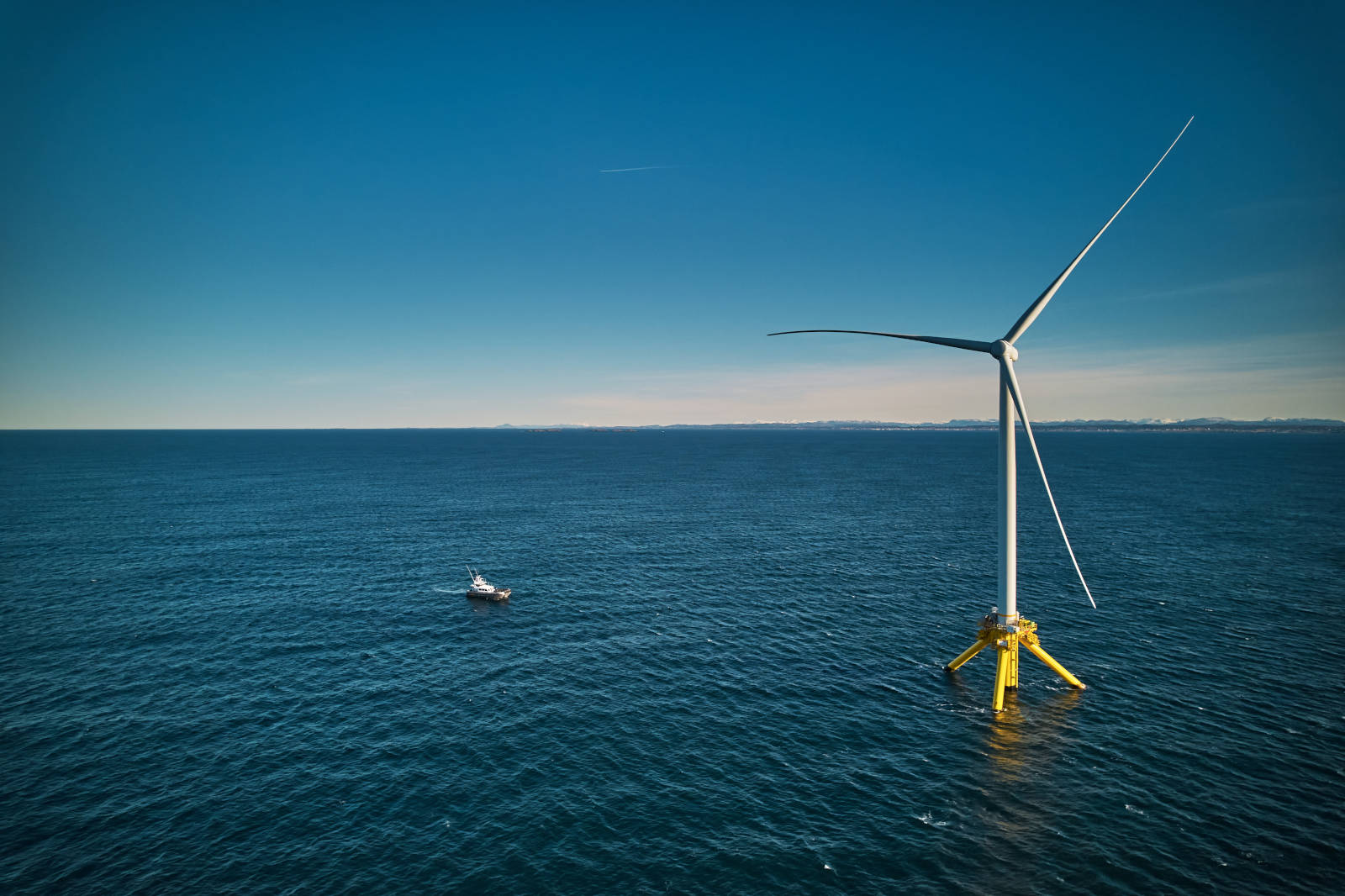 An offshore wind turbine stands tall above the ocean with a service boat nearby under a clear blue sky.