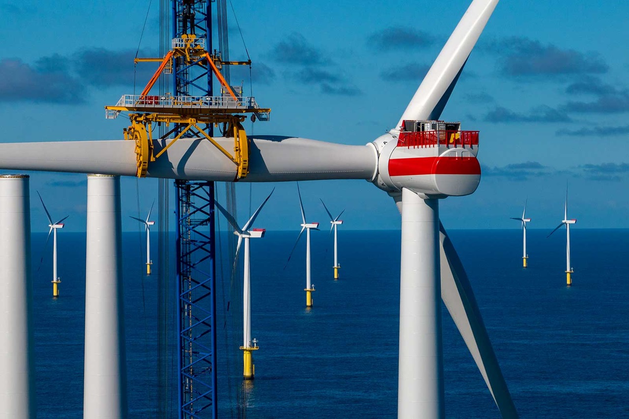 A close-up of a wind turbine under construction at sea, with additional turbines visible in the background against a blue sky.