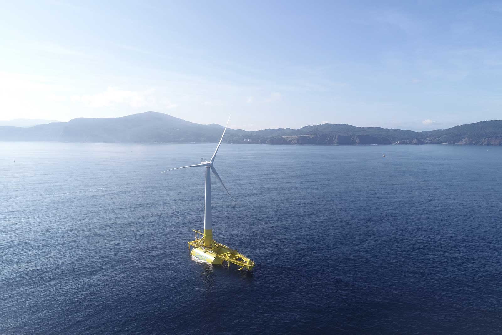 A floating wind turbine on calm waters, surrounded by hills and mountains under a clear sky.