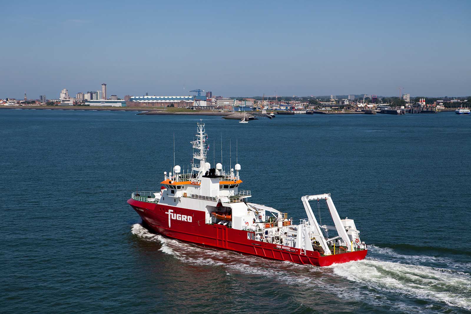 A red research vessel named Fugro sails on calm waters with a city skyline in the background.